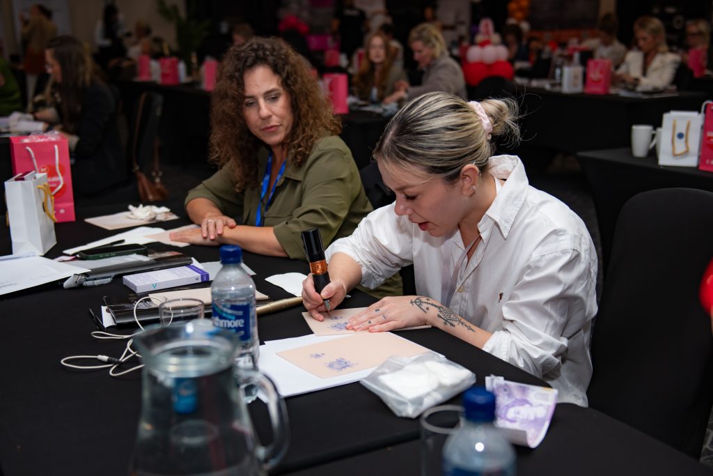 Two artists sitting side by side at a table, drawing brow shapes on practice sheets.