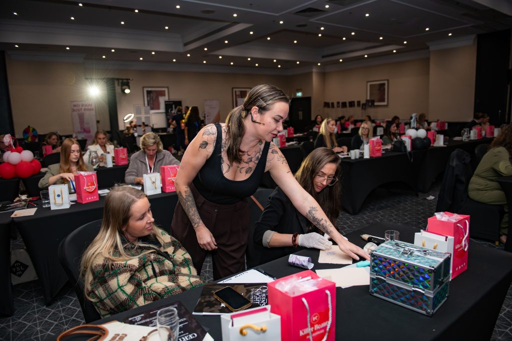 An artist leaning across a table to show a case of products to two seated attendees.