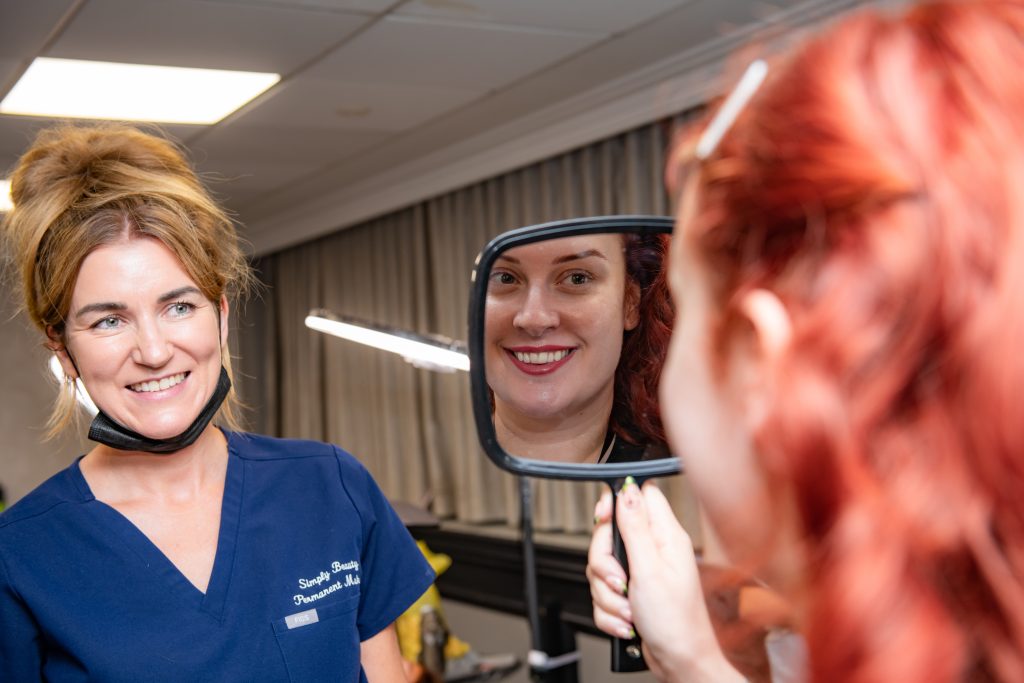 An artist holding up a hand mirror so a client can see her finished brows, both of them smiling.