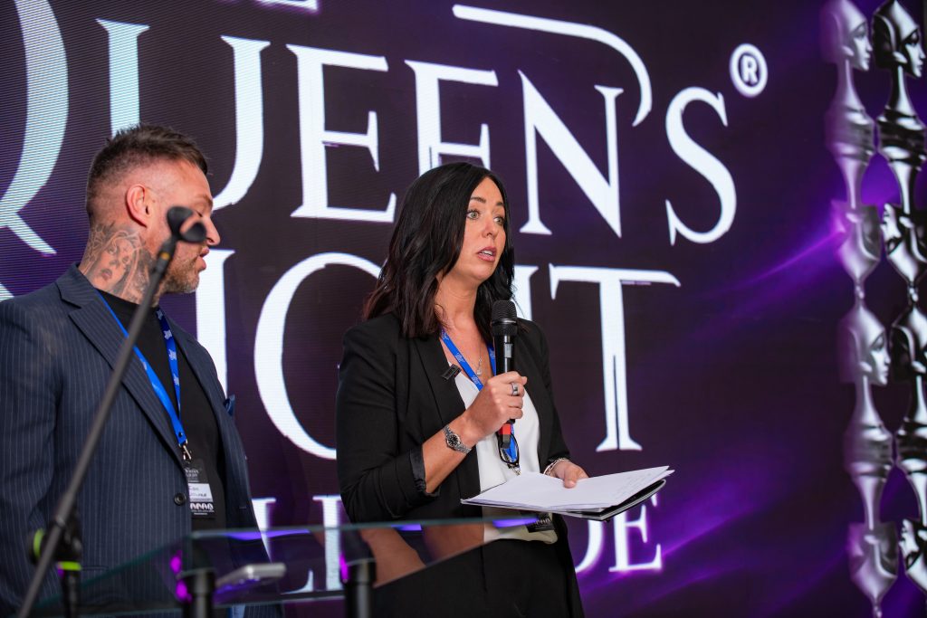 Two people with a microphone in front of the Queen's Night wordmark, 2026.