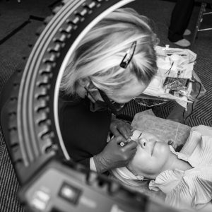 Overhead black and white view through a ring light as an artist in gloves works on the brows of a client lying back on a treatment bed.