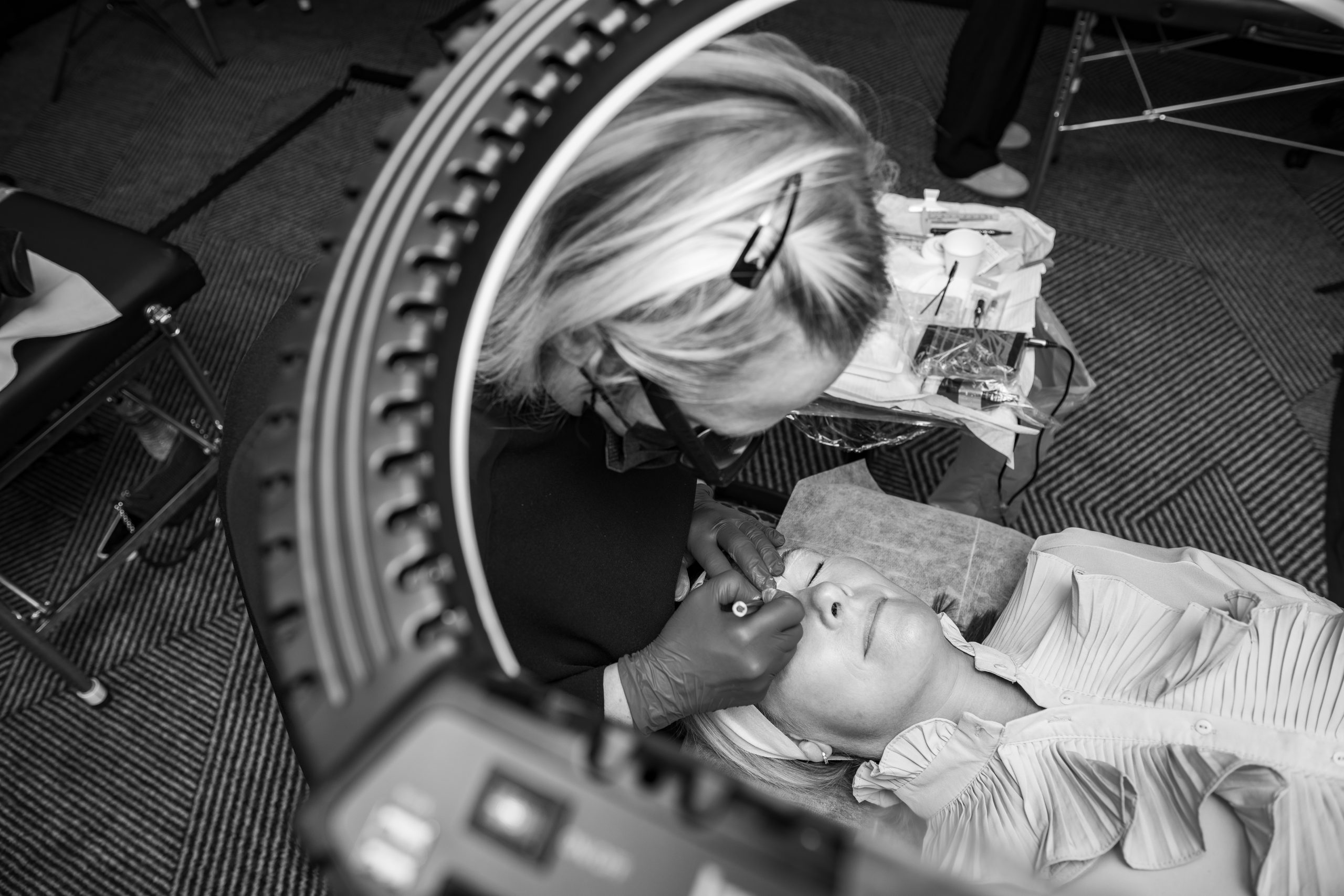 Overhead black and white view through a ring light as an artist in gloves works on the brows of a client lying back on a treatment bed.