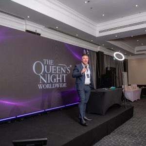 A speaker with a microphone in front of the Queen's Night Worldwide stage screen