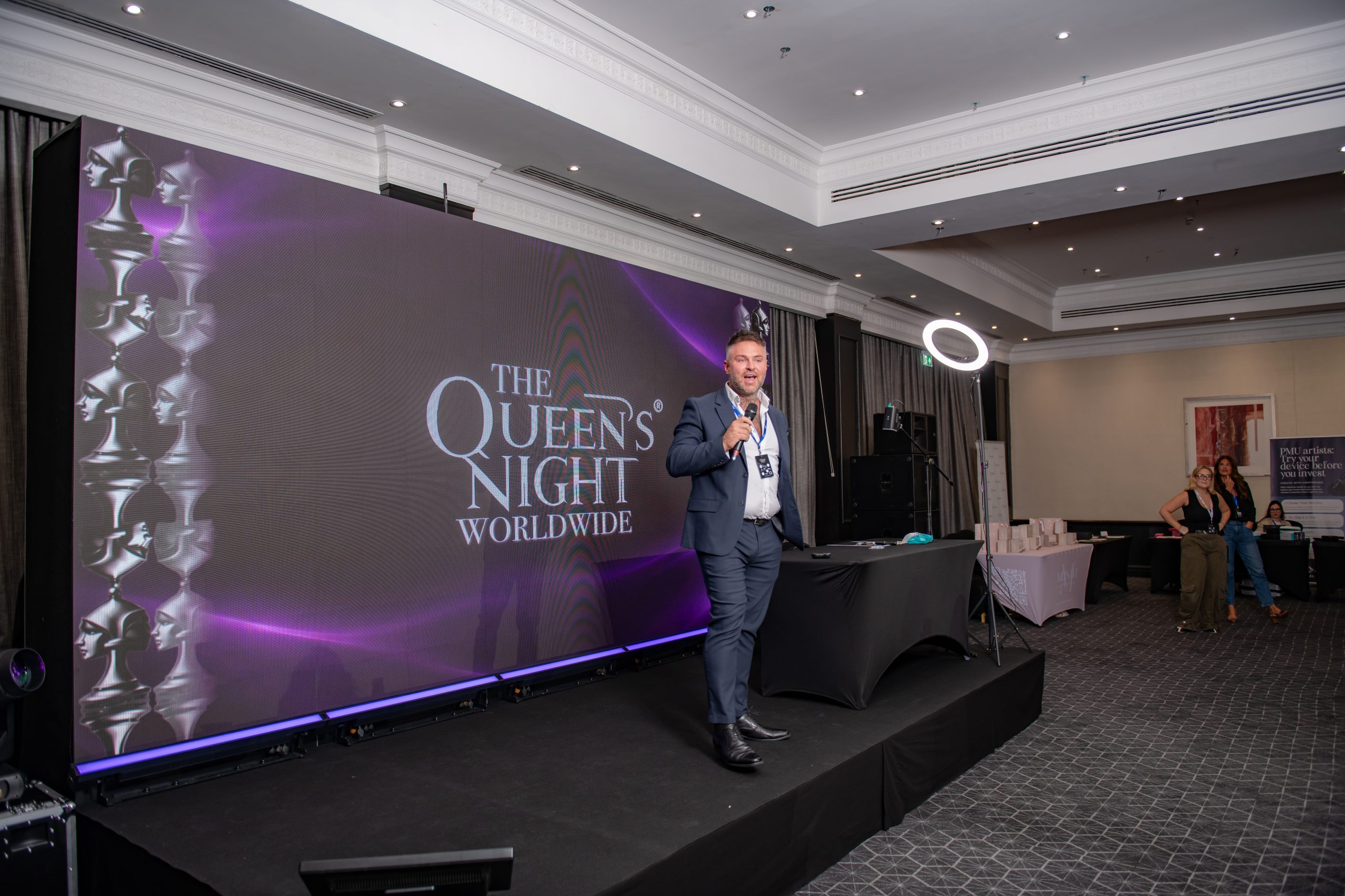 A speaker with a microphone in front of the Queen's Night Worldwide stage screen